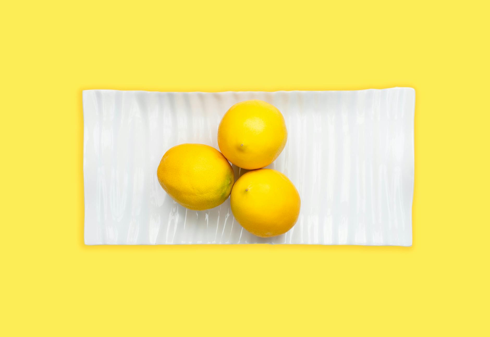 Three fresh lemons on a white plate against a vibrant yellow background
